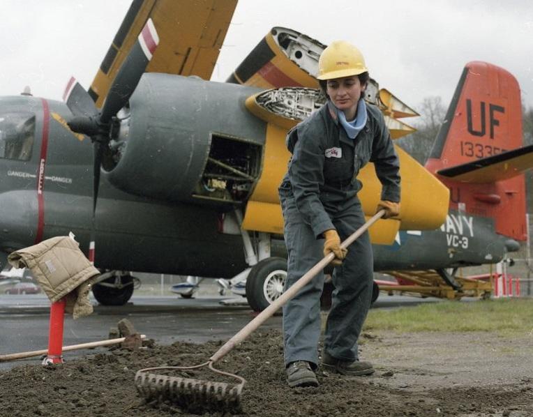 Woman raking dirt in front of a Navy airplane