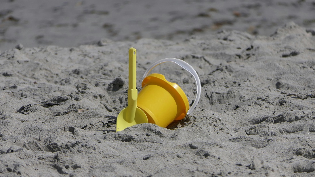 A yellow bucket and shovel lie partially buried in sand on the beach