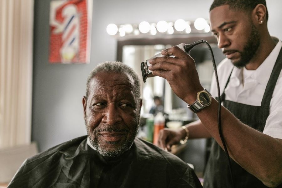 A Black barber with a beard holds a trimmer up to the head of another Black man sitting in a barber's chair