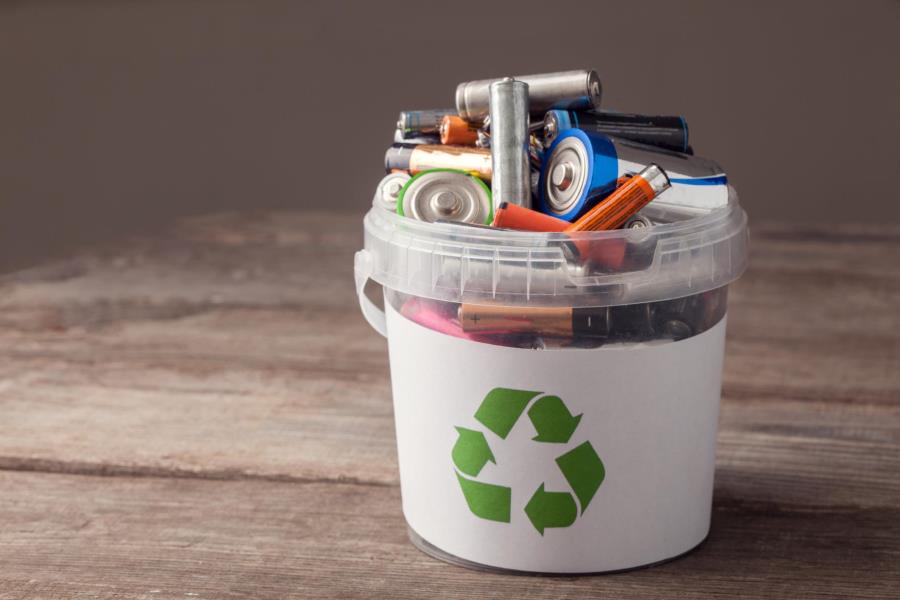 A bucket sitting on a wooden table with the recycling symbol on the front is filled with alkaline batteries