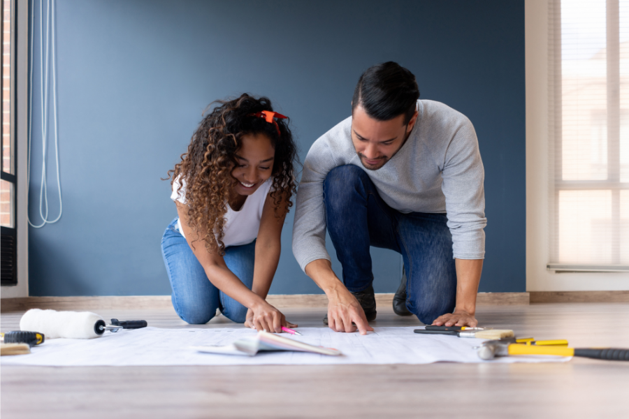 An adult and younger person both wearing light colored shirt and blue jeans are kneeling down in front of a home renovation plan