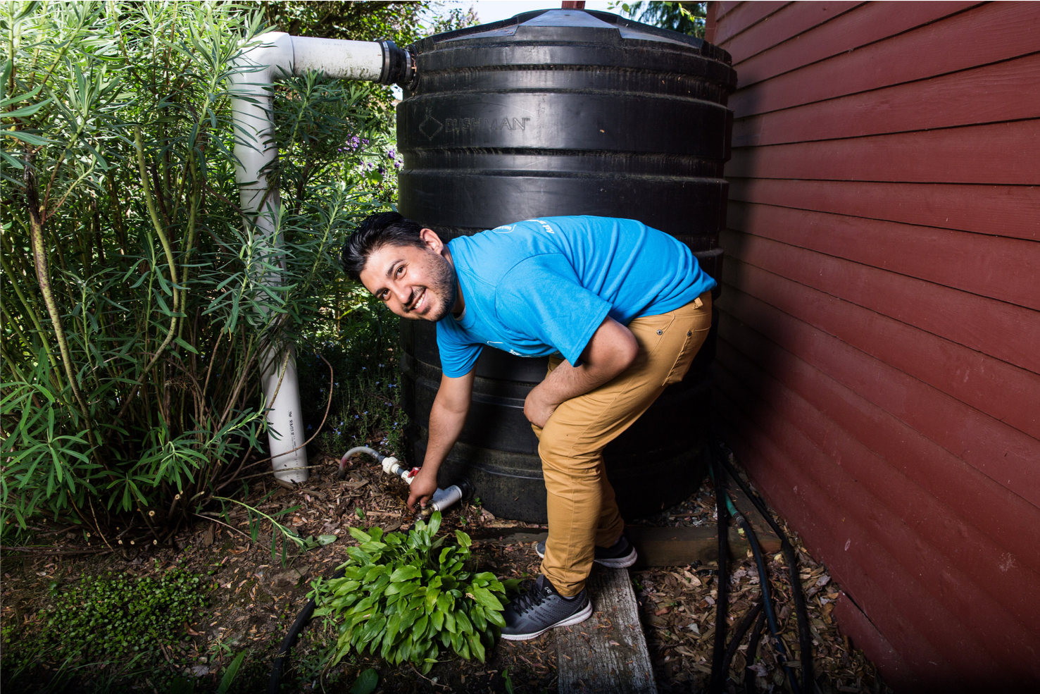 A man in a blue T-shirt checks a valve on a large black water cistern by the side of a red house.