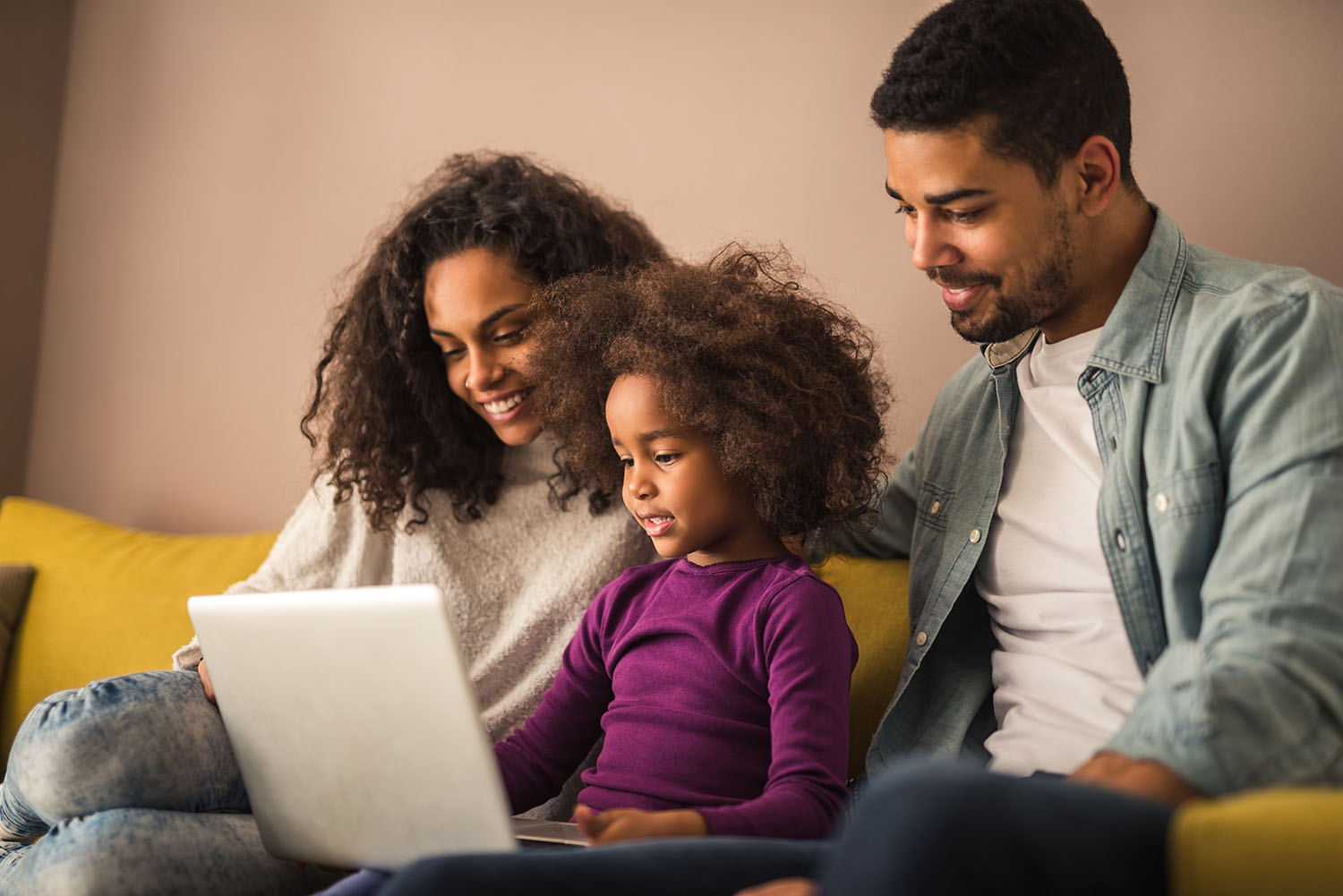 A couple sits with their child on a couch. Everybody is looking at a laptop screen.