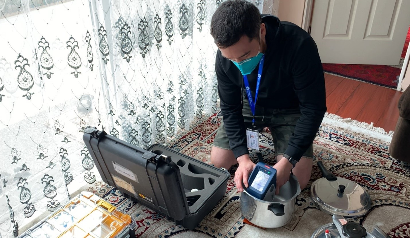 A white man wearing a mask and kneeling on a rug in a home holds a scanning device over a metal pot