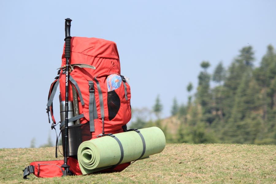 A red backpack with hiking poles and a light-colored sleeping mat on it sits on the ground with trees in the background