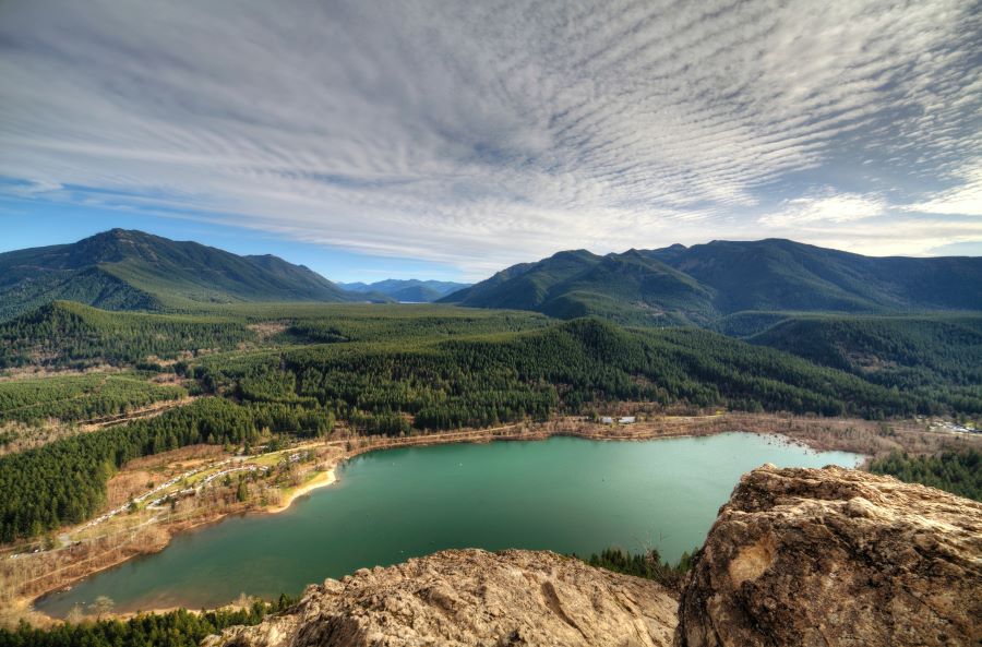  A partially aerial image overlooking Rattlesnake Ledge in Washington of a teal lake surrounded by forests. Tree-covered mountains are in the background.
