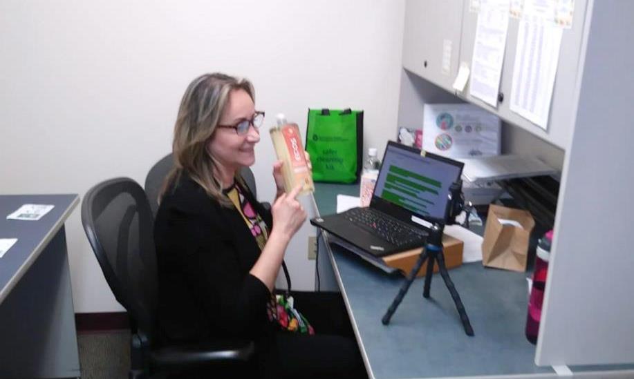 A Sea Mar staff person providing virtual outreach services at their desk. They are holding up a safer product and talking to a camera on a small tripod.
