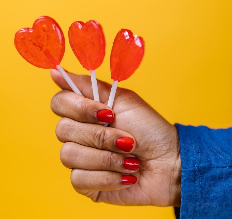 A Black hand with red nails holds out three heart-shaped lollipops in front of a yellow background