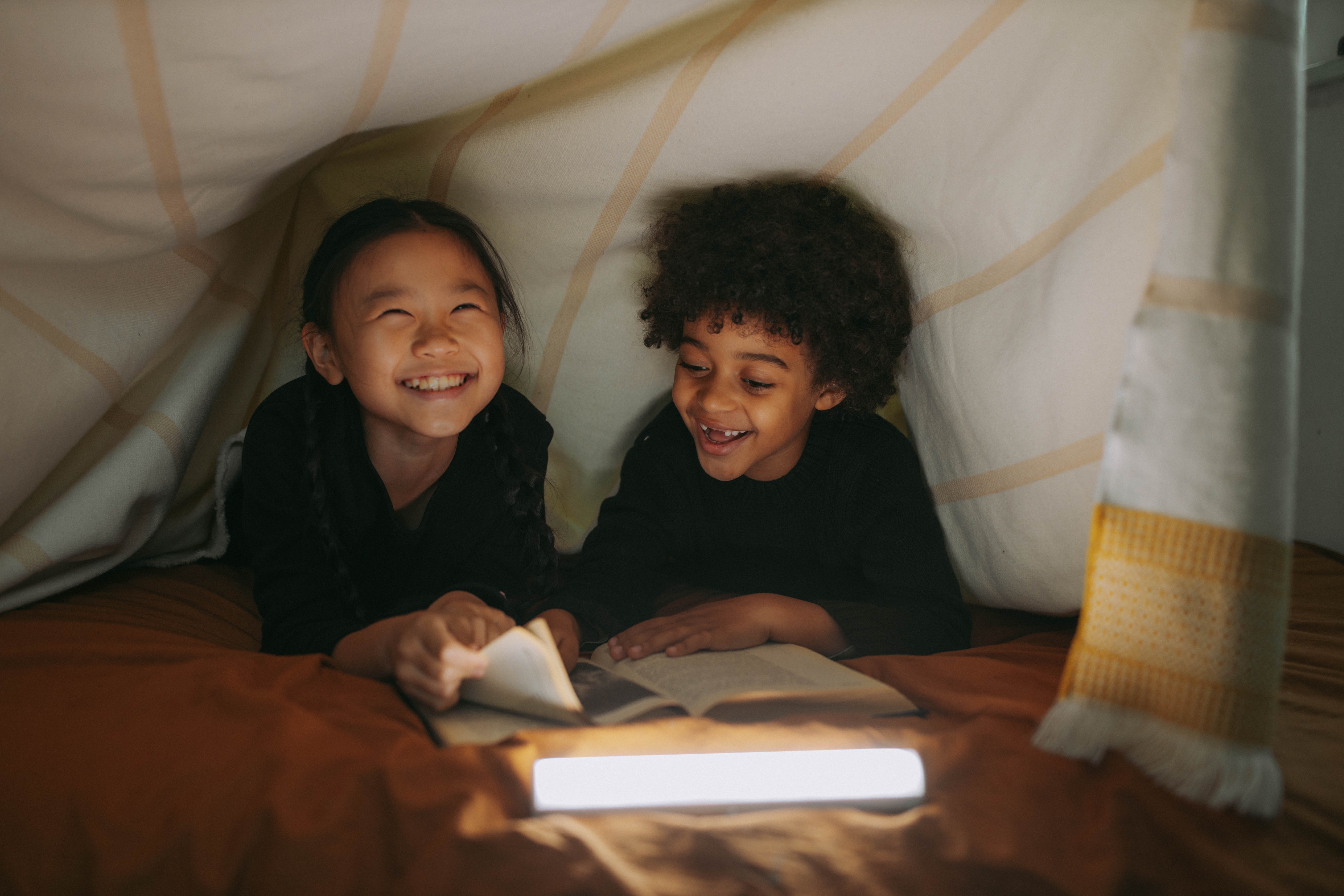 Two laughing children read a book inside a blanket fort.