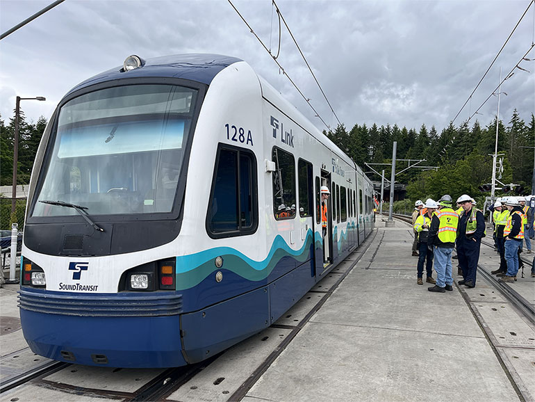 Sound Transit light rail train stopped on the track, with a group of people in hardhats next to it.