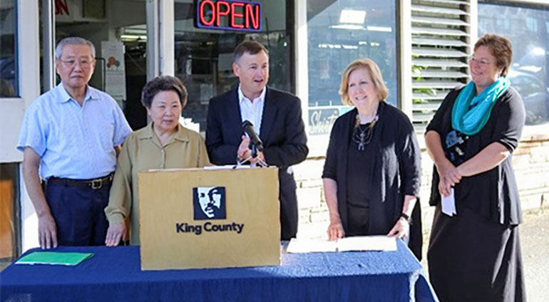 Councilmembers Dembowski and Kohl-Welles with owners of the Sun Drive-In Cleaners in front of a table-top podium.