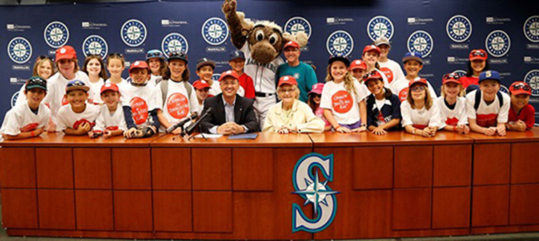 Group of young people and the Mariner Moose mascot behind a counter, with several Mariners logos on the wall and counter front.