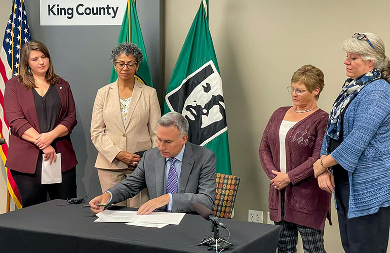 Councilmembers Perry and Balducci and others standing around a desk as Executive Constantine signs the ordinance.