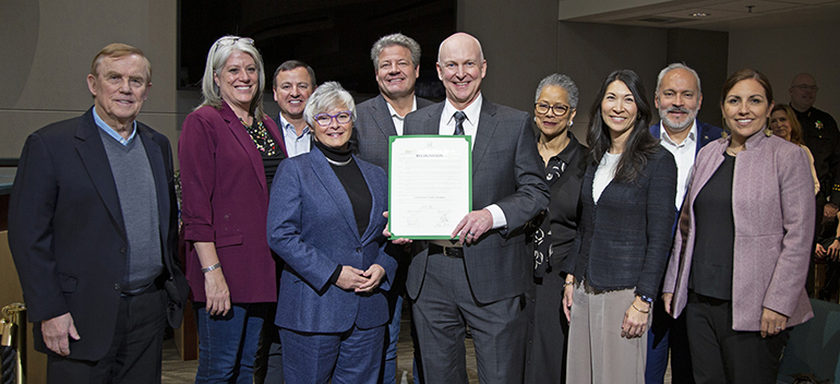 A group of people poses together, with a man in the center holding a document, all facing the camera.