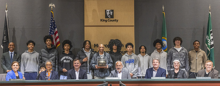 King County Councilmembers with the Rainier Beach Vikings Boys Basketball Team, their coach at the center holding a trophy in the Council Chambers.
