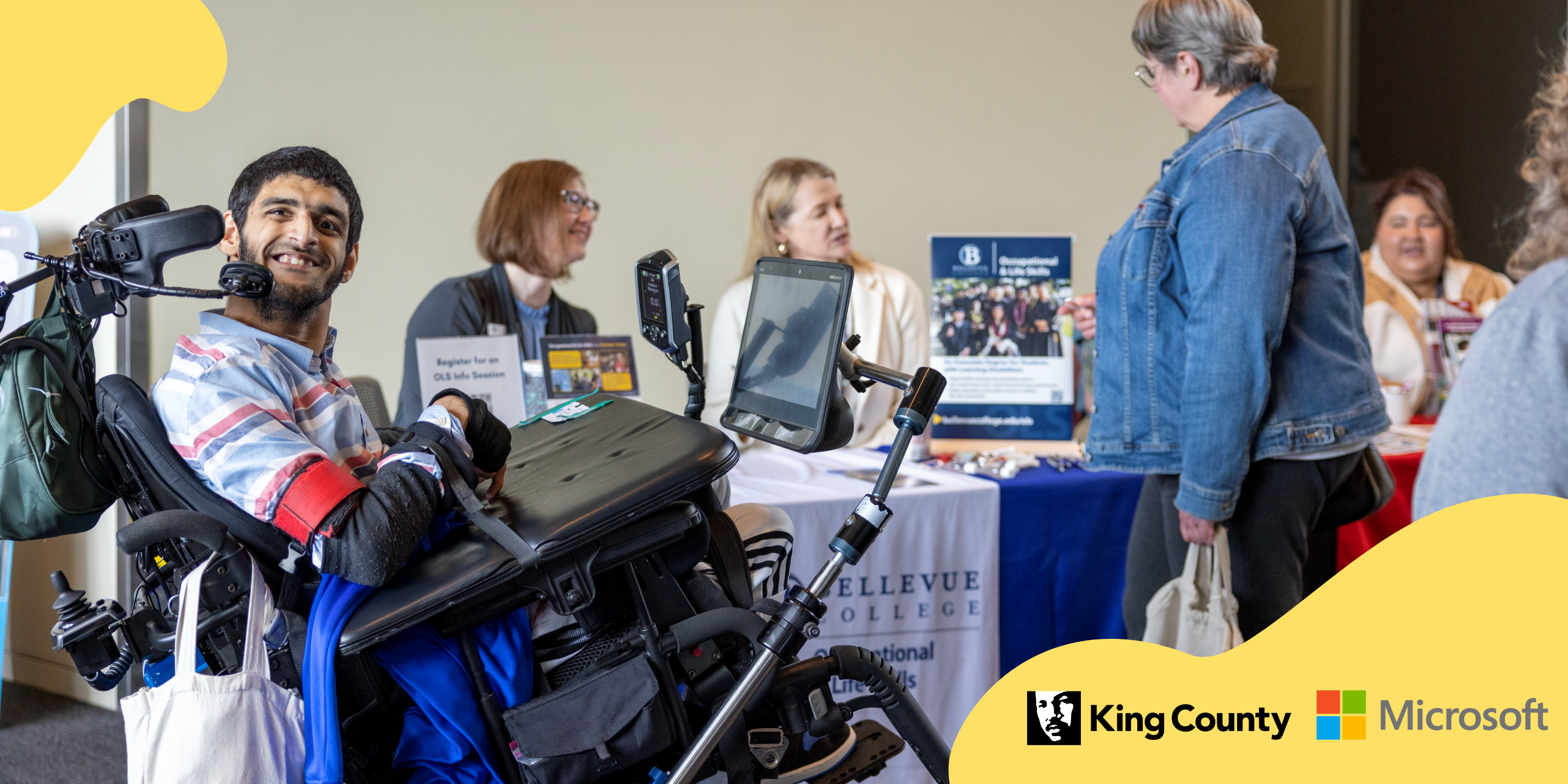 A young man in a wheelchair is smiling at the camera. A resource fair booth is in the background.