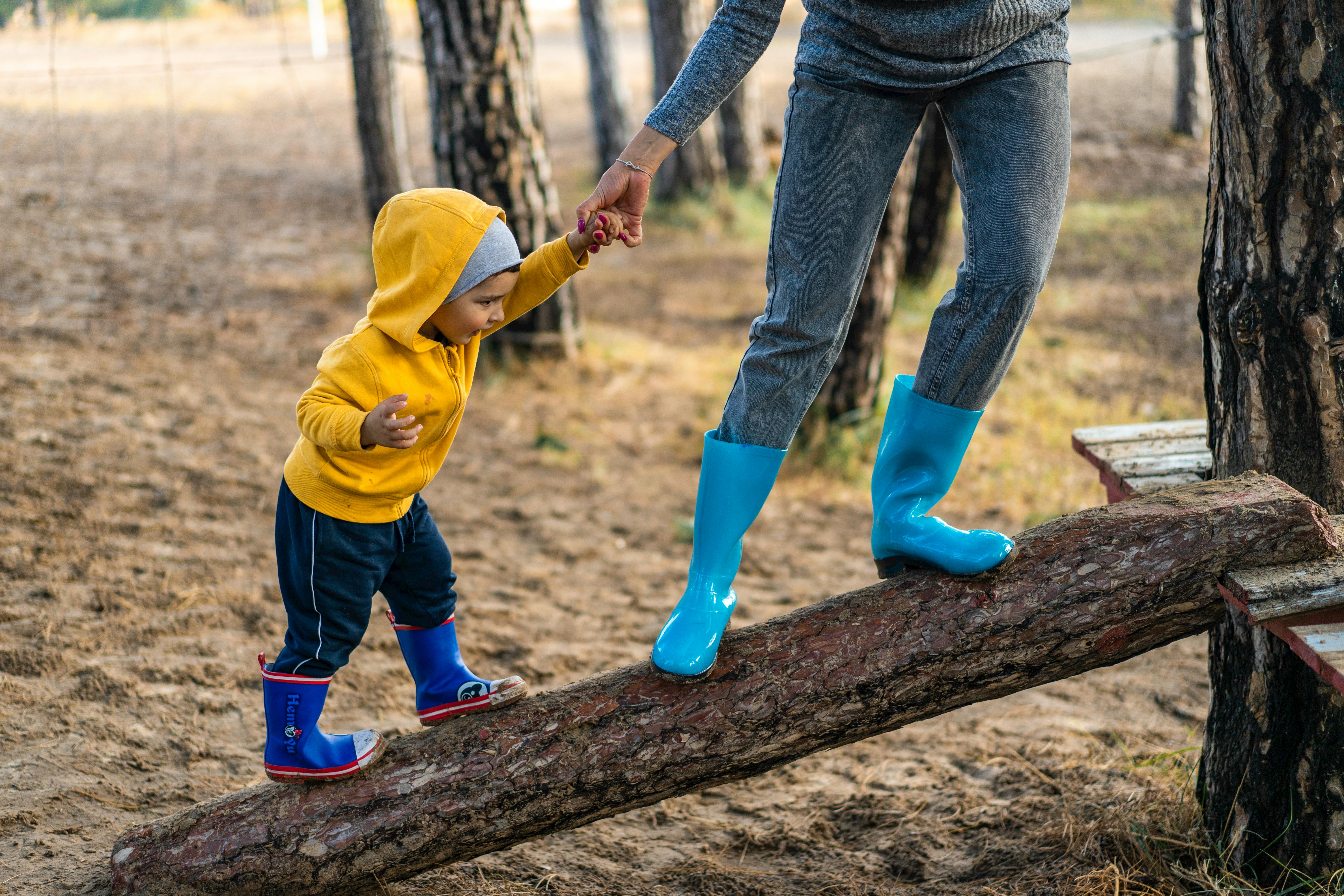 a little boy wearing a yellow jacket and dark blue boots holding hands with an adult wearing bright blue boots and both walking up a fallen tree