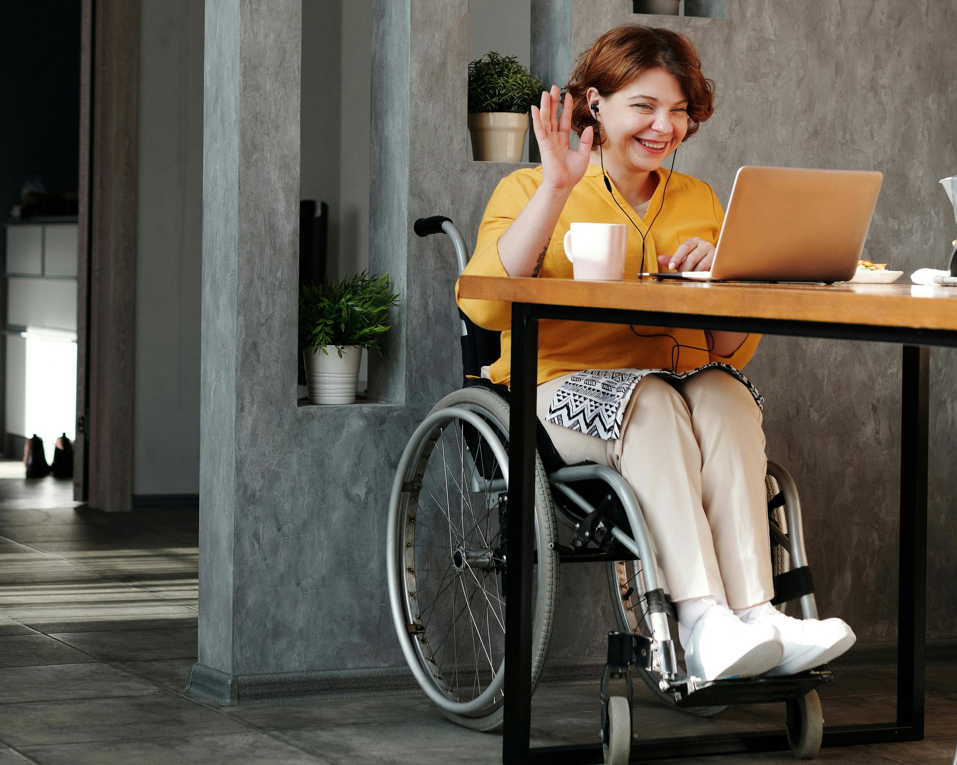A woman in a wheelchair sitting at a table and attending an online meeting on her laptop.