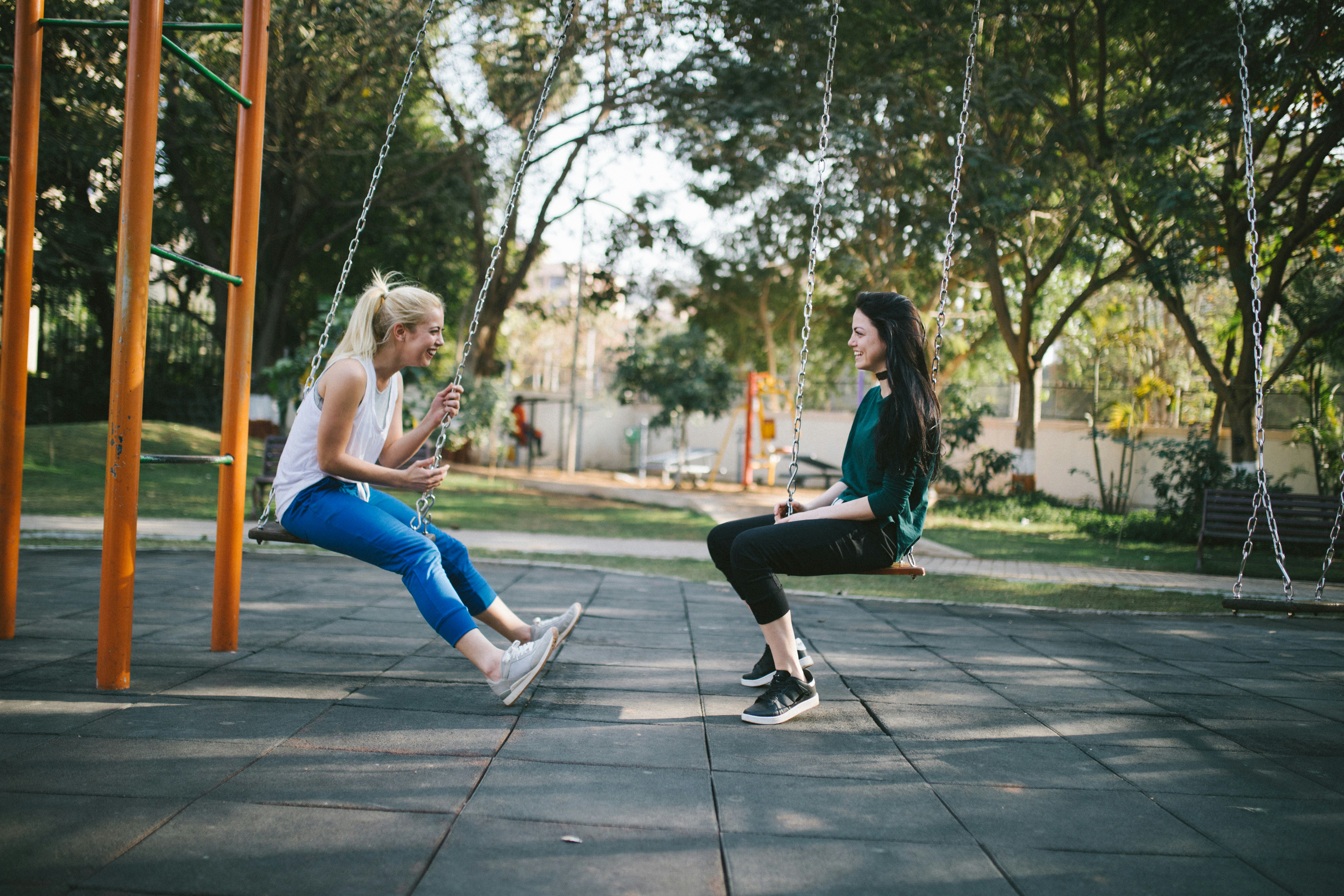 two twenty-something adult women talking to each other, sitting on playground swings