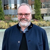 Jay Osborne smiles outside an office building in Downtown Seattle. He is a White man with a gray beard, wearing glasses, and a dark blue jacket.
