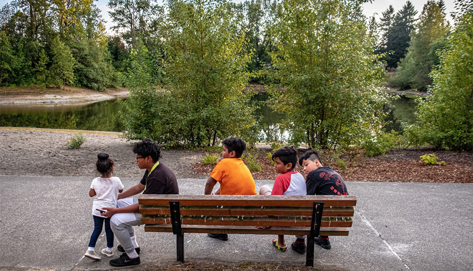 Five children sitting on a park bench near trees and a pond