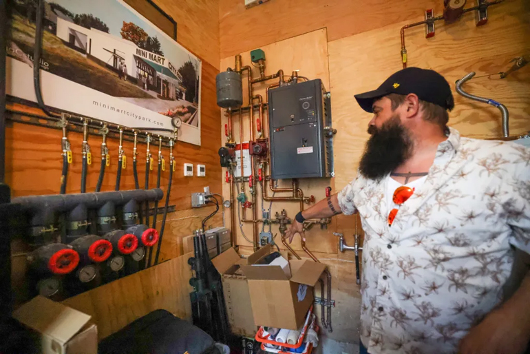 A man with a beard wearing a white collared shirt and ball cap, points to piping and valves that make up a groundwater cleaning system. 