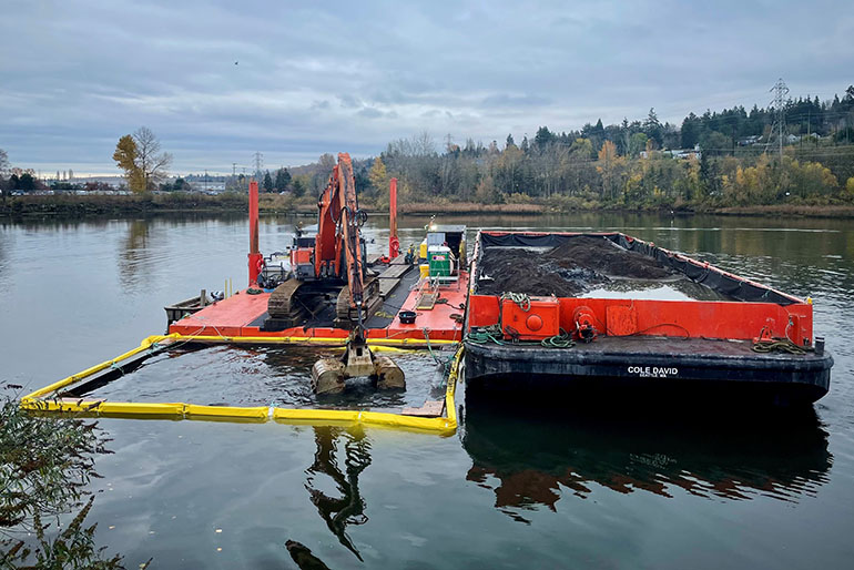 Sediment removal work area out on the water with a digger working within a floating turbidity barrier and a barge to carry away polluted sediment.