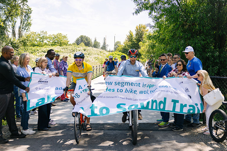 Bikers ride through a breakthrough banner at the opening of the Tukwila segment of the Lake to Sound Trail that will connect Lake Washington at Renton to Puget Sound at the City of Des Moines.