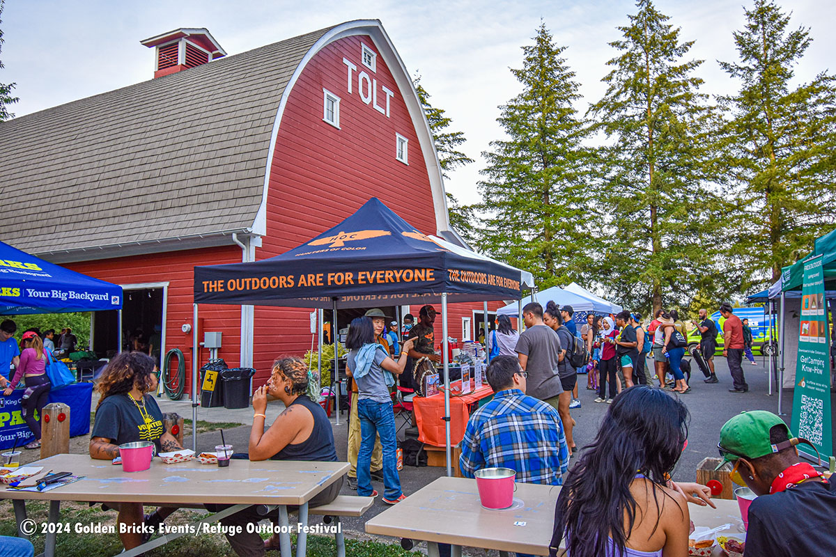 2024 Refuge Outdoor Festival with people eating at tables and visiting displays such as The Outdoors are for Everyone tent at Tolt-MacDonald Park by the big red barn