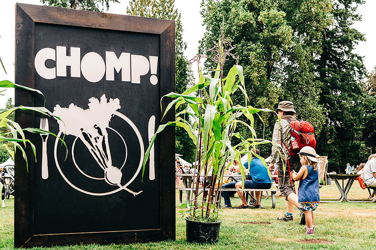 Chomp outdoor sign with cornstalks and a little girl and woman walking by