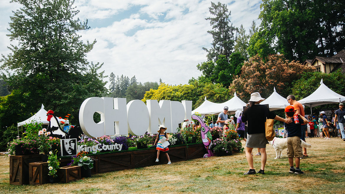 Outdoor sign and flower display on a grassy field at Chomp with people and food tents