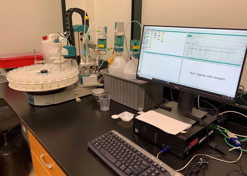A computer monitor sits on a black laboratory bench next to a grey bin with reagent bottles and an auto-titrator instrument sample carousel. This instrument is used to measure alkalinity. The screen shows data from a sample analysis.