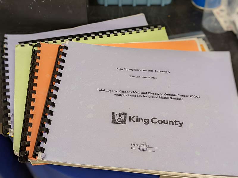 Four comb-bound logbooks with yellow, orange, and white covers are fanned out on a black laboratory bench.