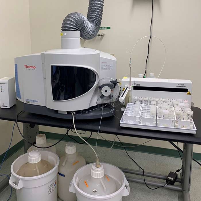 An Inductively Coupled Plasma (ICP) instrument and autosampler sit on a table with waste containers underneath and sample vials in white holders to the side.
