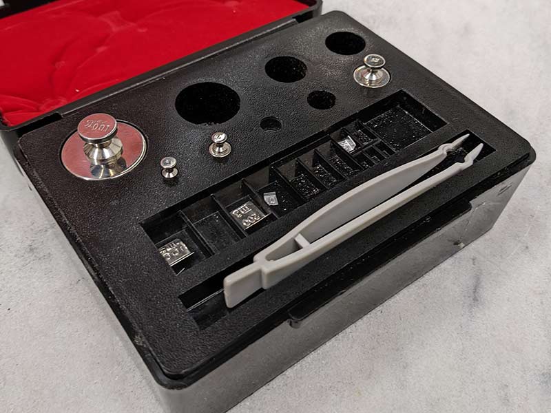 A set of certified, silver-colored weights used for balance calibration are in a black storage container with red lining in the lid on a marble tabletop