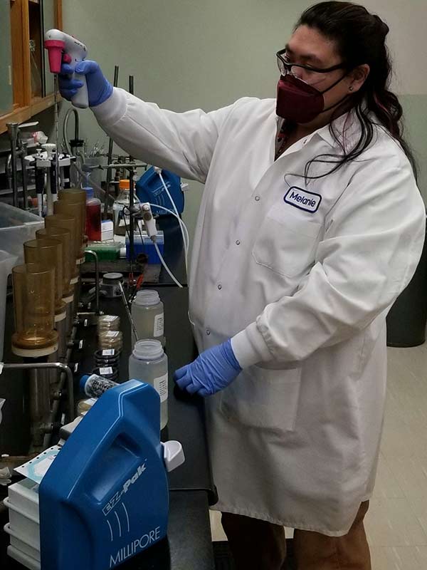 A scientist wearing a white laboratory coat and blue gloves gently places a white filter onto an agar plate.