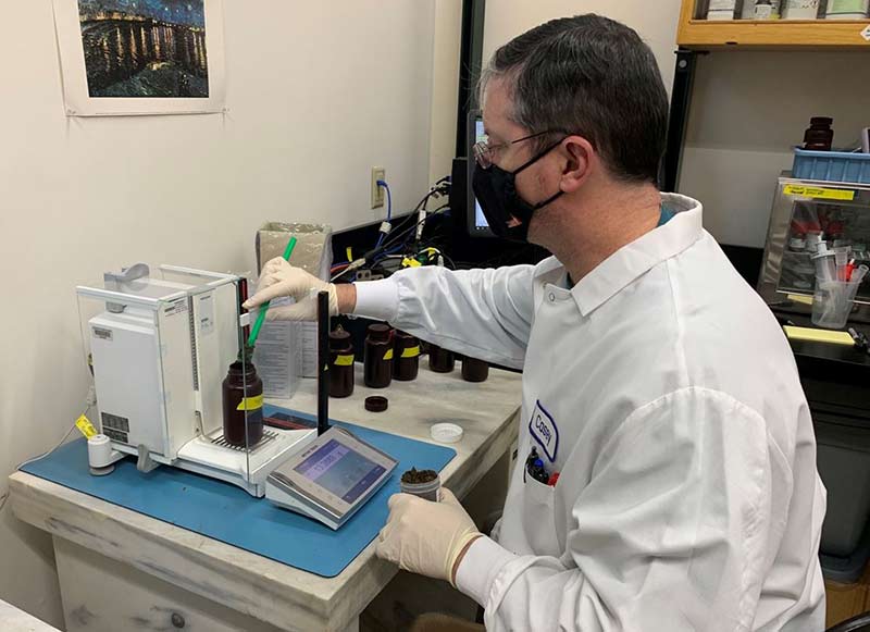 A scientist wearing a white laboratory coat and gloves adds dark colored material to an amber plastic bottle on a balance in the laboratory.