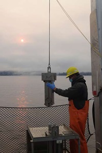 A scientist wearing a PFD, orange waterproof pants, and a yellow hardhat steadies a sediment collection instrument on the rear deck of the R/V SoundGuardian with water, trees, and a hazy sun in the background.
