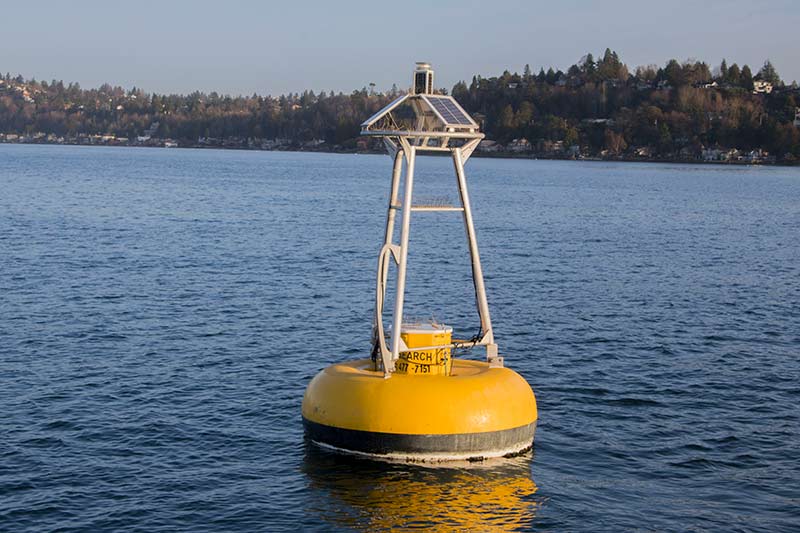 A yellow, wide-disk shaped buoy with a grey metal tripod frame and scientific instruments on top of it floats in water with trees on a hill in the background.