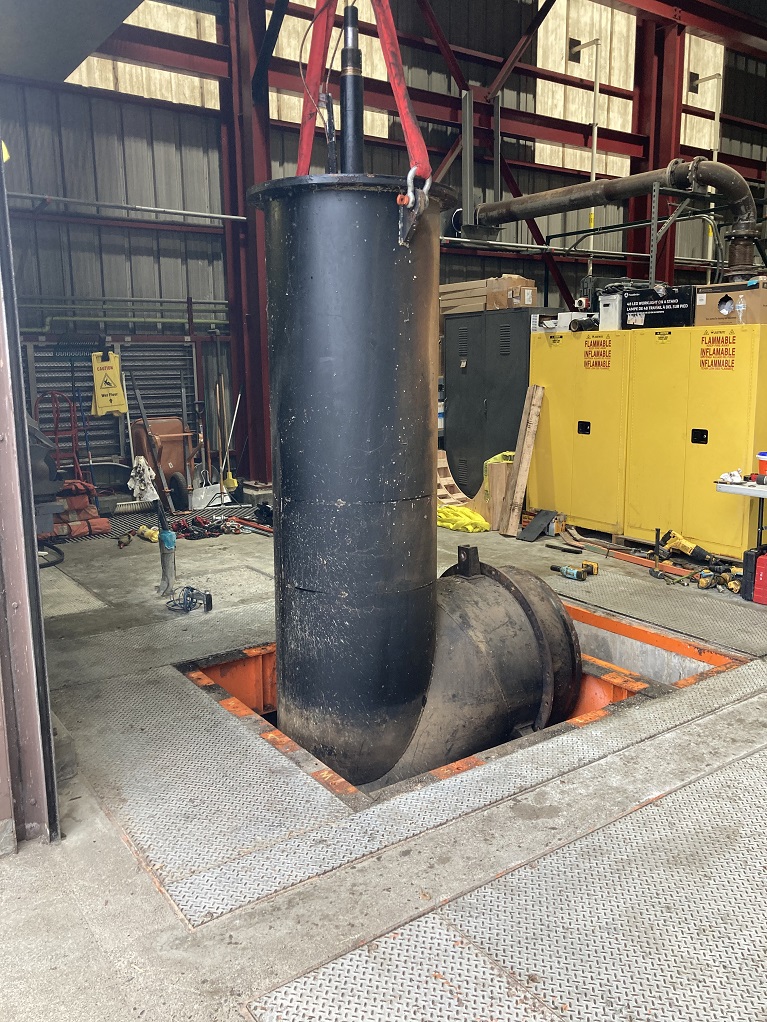 Large black pump part being lifted by straps out of a ground inside a building.