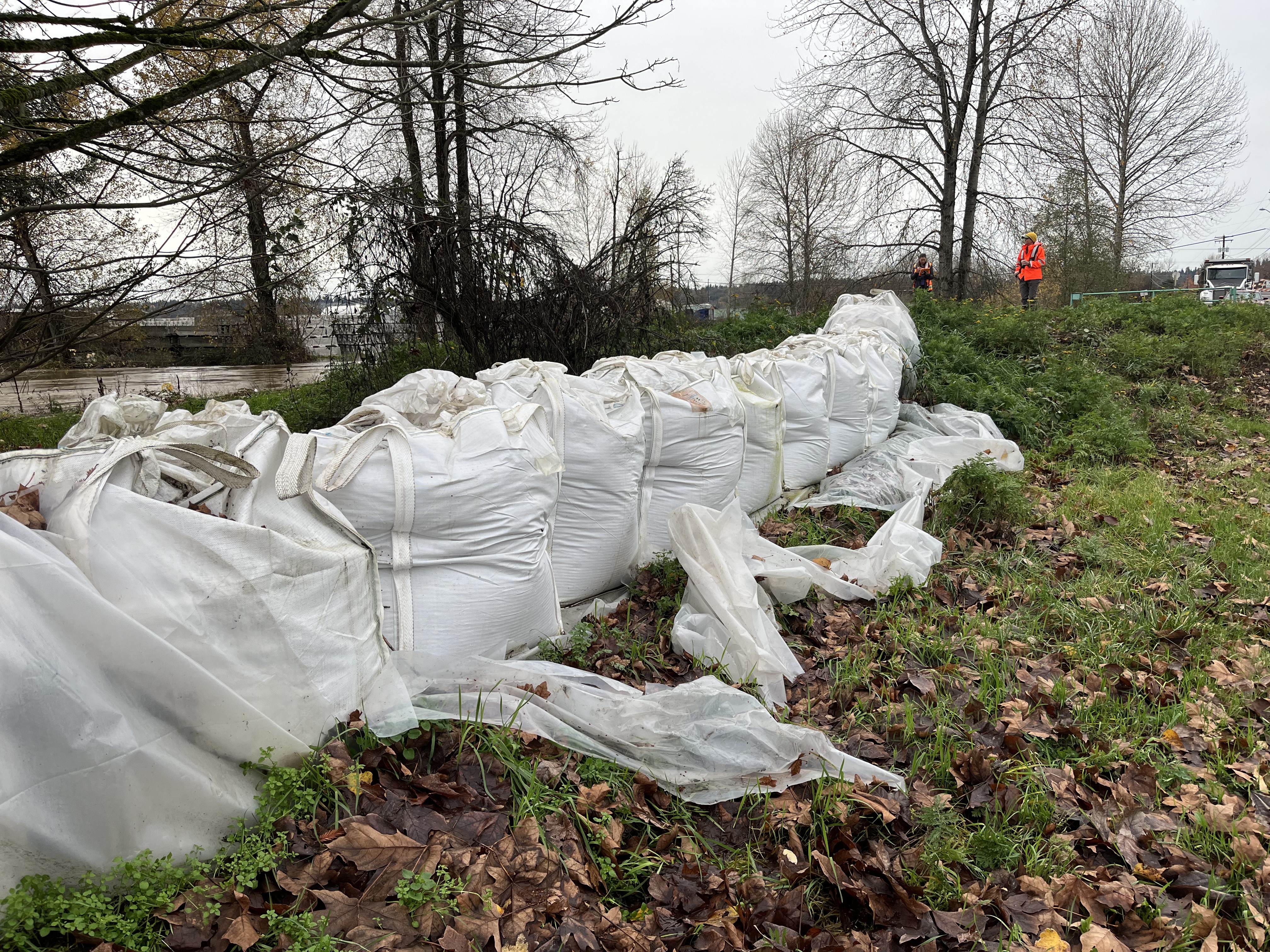 Large white sacks of sand line a riverbank near the Green River. Two people in orange and black safety gear take flood observations.