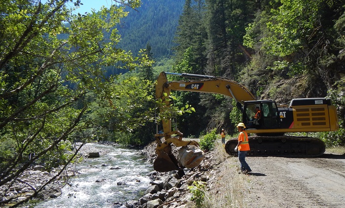 An excavator on a dirt road digs into rocks on a riverbank while a construction worker is in the foreground.