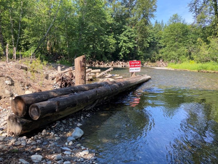 Stacked logs lay on a riverbank on a sunny day.
