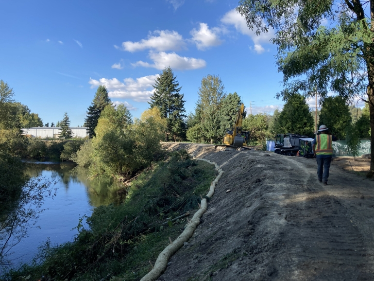 A person in a high visibility vest and hard hat walks on the dirt-covered McCoy Levee near construction equipment. Blue sky is reflected in the Green River next to the levee.