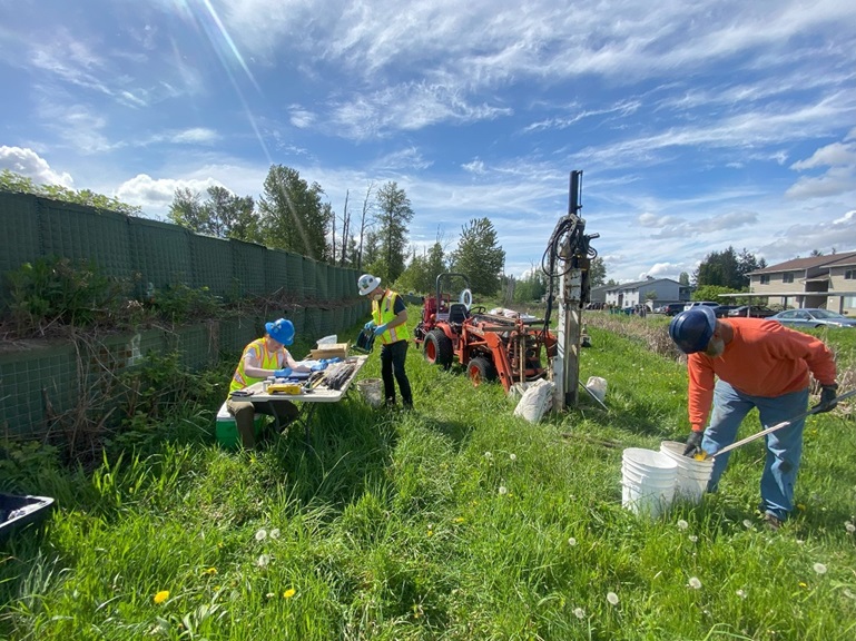 A photo that shows a dumpsite sampling investigation happening in Pacific City Park. Two people in bright yellow utility vests are working with soil samples and instruments at a temporary table. A person in the foreground is digging up some soil with shovel, and putting that soil into a white bucket.