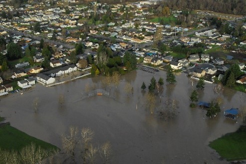 A photo showing an aerial view of Pacific City Park during or after a heavy flood. Water covers the majority of the open space in the park and extends toward the road. Some water appears to be flanking apartments along the road.