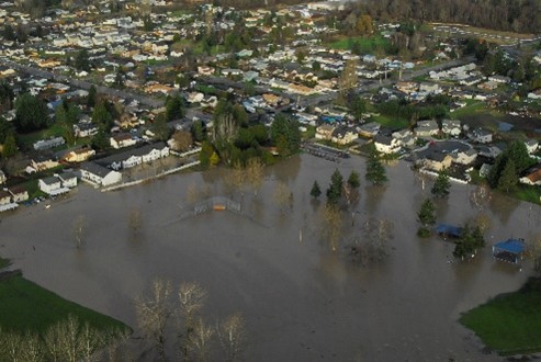 A photo showing an aerial view of Pacific City Park during or after a heavy flood. Water covers the majority of the open space in the park and extends toward the road. Some water appears to be flanking apartments along the road. 