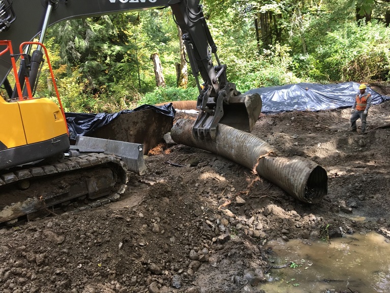 Yellow excavator grabs a metal tube while a construction worker watches.
