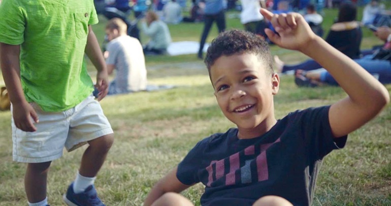 Boy sitting in a grassy field raising his hand.