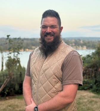 A man with a beard standing in front of a lake